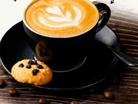 A cappuccino with latte art in a black cup, with a chocolate chip cookie on the saucer