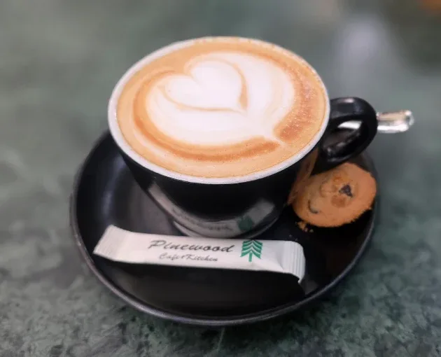 A coffee with a heart in the milk foam, a cookie and a Pinewood sugar sachet