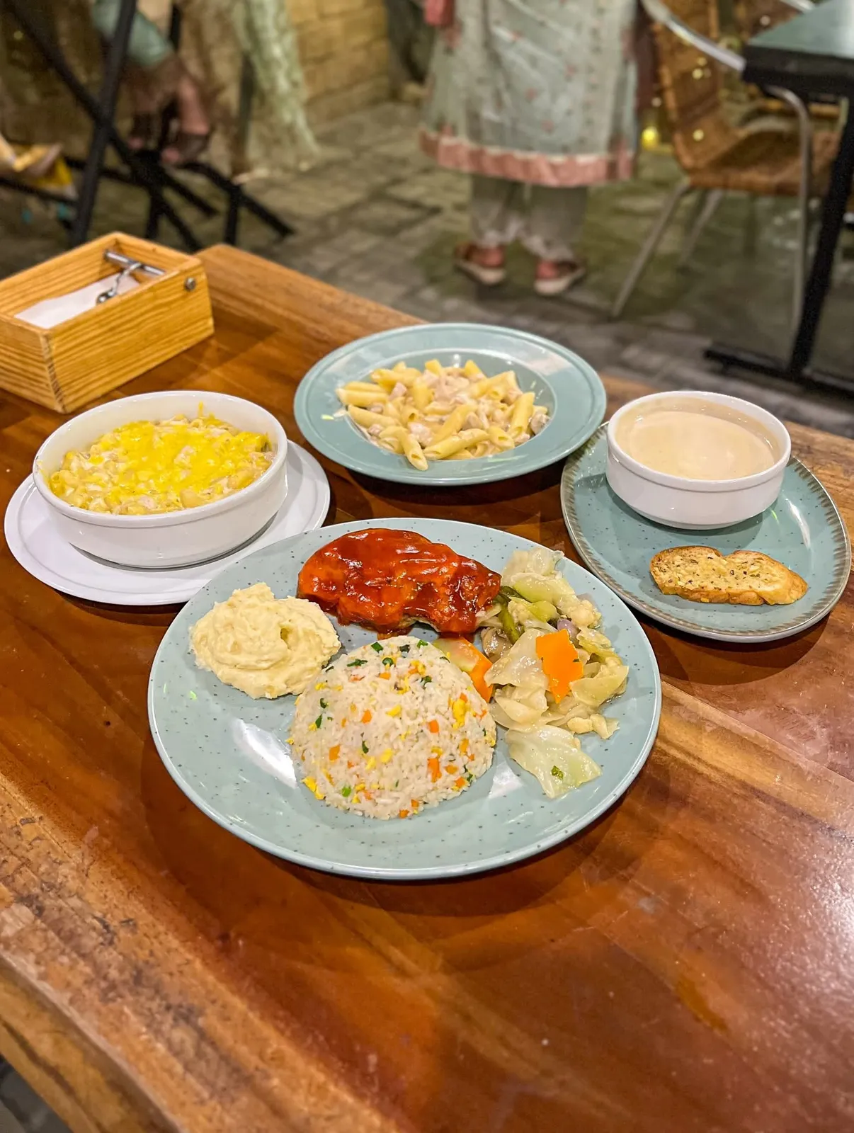 A chicken plate with fried rice, a bowl of mac and cheese, penne pasta and soup on a wooden table